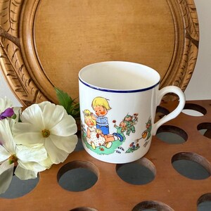May include: White ceramic mug with a blue rim and handle, featuring a colourful illustration of children and gnomes. The mug is set against a wooden background with a decorative floral arrangement.