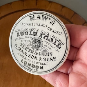 May include: A white ceramic lid for a jar of tooth paste. The lid has black text that reads "MAW'S INDIAN BETEL NUT OR ARECA TOOTH PASTE UNEQUALLED FOR EFFECTS ITS SALUTARY ON THE TEETH & GUMS S. MAW, SON & SONS ALDERSGATE STREET LONDON ENGLAND".