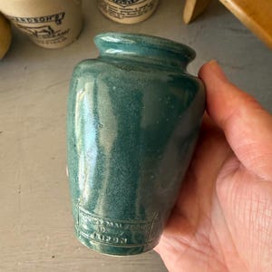 May include: A small, teal-colored ceramic jar with a smooth, glossy finish and a narrow neck. The base of the jar features embossed text that reads "MACKEY MALZEARD" and "RIPON" enclosed within a rectangular border design on the front surface.