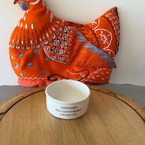 May include: An orange chicken-shaped tea cosy with blue and white embroidered details. A small white ceramic bowl with the text "SHIPPAMS HIGH CLASS POTTED MEAT CHICHESTER" sits on a wooden tray.