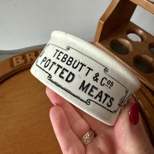 May include: A small, white ceramic dish with the text "TEBBUTT & CO'S POTTED MEATS" in black lettering. The dish has a curved shape and is held in a hand. A wooden rack and a wooden cutting board are in the background.