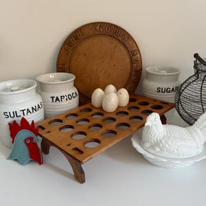 May include: A wooden egg rack with 24 slots, a white ceramic hen, and four white eggs. There are three white ceramic jars with black lettering that say "SULTANAS", "TAPIOCA", and "SUGAR". A wooden round chopping board with the words "Come and Go" carved into it.