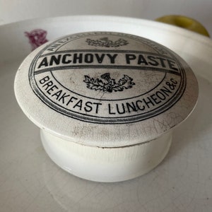 May include: White ceramic jar with a lid. The lid has a black and white design that says "An Excellent Anchovy Paste Breakfast Luncheon &c".