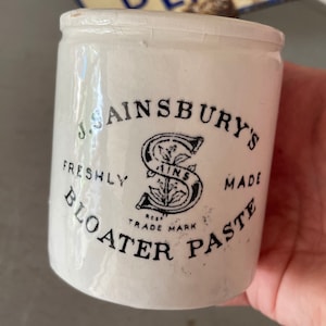 May include: A white ceramic jar with a black label that reads "J. Sainsbury's Freshly Made Bloater Paste" and features a stylised "S" with a floral design. The label also includes the text "Regd Trade Mark".