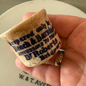 May include: A small, cylindrical ceramic pot with a cream-coloured, crackled glaze and dark blue printed text that reads: Prepared only by Beach & Barnicott, successors to the late Dr. Roberts, Bridport. The pot is held against a white surface.