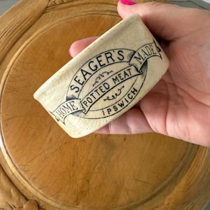 Peut inclure: Un pot en céramique blanche avec une inscription noire qui dit "Seager's Home Made Potted Meat Ipswich". Le pot est décoré d'une bannière noire en ruban.