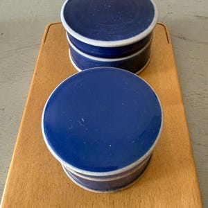 May include: Two round, deep blue ceramic containers with matching lids, each featuring a white rim. The containers are positioned on a rectangular wooden board that has a small, decorative butterfly-shaped handle at the top edge.