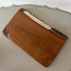 May include: A wooden cheese board with a stainless steel knife. The board is rectangular with a curved edge and floral carving. The knife has a wooden handle with a floral design. The board and knife are brown.