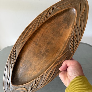 May include: An oval wooden tray with a smooth, dark brown centre and a carved border featuring a repeating leaf and vine pattern. The wood grain is visible throughout the surface, and the edges are defined by intricate, textured relief carvings.