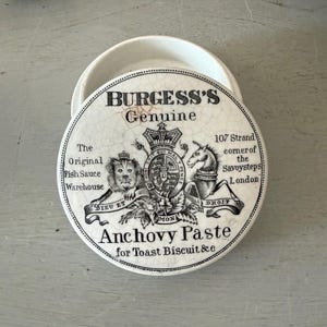 May include: A round, white ceramic lidded jar featuring black text and a heraldic crest. The lid reads Burgess's Genuine Anchovy Paste for Toast Biscuit & c, with illustrations of a lion and a unicorn flanking a central shield and crown emblem.
