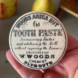 May include: A white ceramic jar lid with black lettering that reads "WOODS ARECA NUT 6d TOOTH PASTE Forremoving Tartor and whitening the tieth withent injuring the Enumer PROPRIETOR W.WOODS CHEMIST PLYMOUTH".