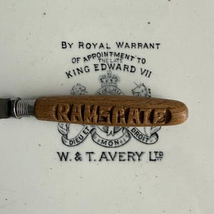 May include: A butter knife with a wooden handle, engraved with the name "RAMSGATE". The knife has a silver blade and is set against a white background with text that reads "By Royal Warrant of Appointment to King Edward VII".