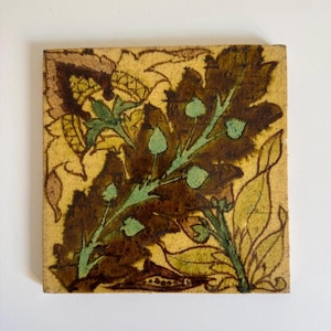 May include: Square ceramic tile with a botanical design. The tile features a central brown leaf with green accents and a yellow background. Other leaves and floral elements are also visible.