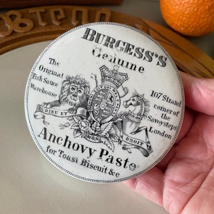 May include: A white ceramic jar lid with black lettering and a crest. The lid reads "Burgess's Genuine The Original Fish Sauce Warehouse 107 Strand corner of the Savoy steps London Anchovy Paste for Toast Biscuit &c".