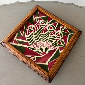 May include: A framed artwork with a geometric design in red, green, and cream colours. The artwork is encased in a square, wooden frame with a decorative edge. The design features stylised shapes and patterns.