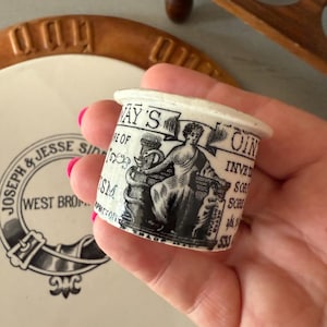 May include: A small, cylindrical ceramic container with a white base and a black and white illustration. The illustration features a classical figure and text that reads "WAY'S RE OF ISM". The container is held in a hand.
