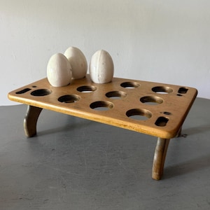 May include: A wooden egg holder with three white eggs. The holder is rectangular with rounded holes and curved legs. The wood has a warm, natural tone, and the eggs are smooth and oval-shaped.