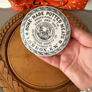 May include: A circular, off-white container with black text reading "HOME MADE POTTED MEATS" and "MANUFACTURED BY THE ARMY AND NAVY CO-OPERATIVE SOCIETY LTD". The container is held in a hand, with a wooden tray in the background.