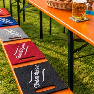 Può includere: Una serie di cuscini rettangolari in feltro rosso, blu, grigio, rosso scuro e nero, ciascuno dotato di una maniglia ritagliata e della scritta bianca "Bierbank-Rutscherl" stampata, disposti lungo una panca da birra in legno all'aperto.