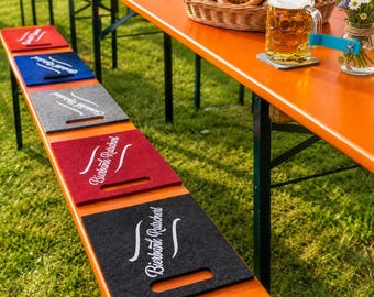Puede incluir: Una serie de cojines rectangulares de fieltro en rojo, azul, gris, rojo oscuro y negro, cada uno con un asa recortada y el texto blanco "Bierbank-Rutscherl" impreso, dispuestos a lo largo de un banco de cerveza de madera al aire libre.