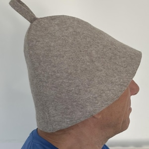 Könnte beinhalten: Ein hellgrauer Sauna-Hut mit einer Schlaufe oben. Der Hut besteht aus einem filzartigen Material und hat eine Glockenform. Er wurde entwickelt, um in einer Sauna getragen zu werden, um den Kopf vor Hitze zu schützen.