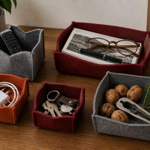 Può includere: Un set di cinque vassoi portaoggetti in feltro grigio, rosso scuro e arancione. I vassoi contengono vari oggetti tra cui un telecomando, un caricabatterie bianco, chiavi, occhiali, una penna, noci e uno schiaccianoci su una superficie in legno.
