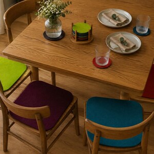 Puede incluir: Una mesa de comedor de madera con cuatro sillas, cada una con un coj&iacute;n de asiento cuadrado de fieltro en verde lima, morado oscuro, azul cerceta y rojo. La mesa tiene un florero de vidrio, un soporte para posavasos, dos platos y dos vasos.