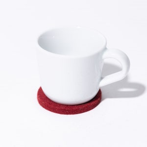 Puede incluir: Una taza de café de cerámica blanca con asa sobre un posavasos de fieltro rojo.