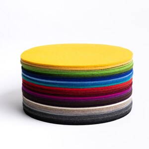Puede incluir: Una pila de posavasos circulares de fieltro en una variedad de colores que incluyen amarillo, verde, azul, rojo, morado, marr&oacute;n y tonos de gris. Los posavasos est&aacute;n apilados, creando una forma cil&iacute;ndrica con una superficie superior plana.