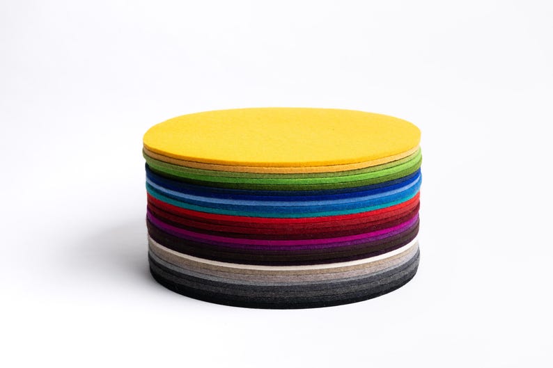 Puede incluir: Una pila de posavasos circulares de fieltro en una variedad de colores que incluyen amarillo, verde, azul, rojo, morado, marr&oacute;n y tonos de gris. Los posavasos est&aacute;n apilados, creando una forma cil&iacute;ndrica con una superficie superior plana.