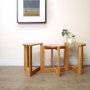 May include: Three wooden side tables in different shapes: a semi-circle, a circle, and a rectangle. The tables are made of light-coloured wood and have a simple, modern design. A framed picture hangs on the wall above the tables. A small vase with plants and a mug are on the rectangular table.