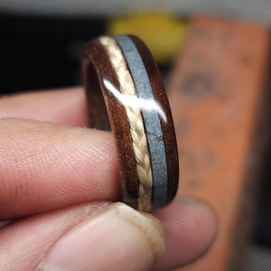 May include: A wooden ring featuring a braided central band and a grey inlay. The ring's outer section is a dark brown, and the braided part is a light beige. The ring is held by a person's fingers.
