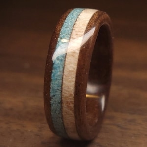 May include: A wooden ring with a dark brown exterior, featuring a turquoise and light wood inlay. The ring's design includes a central turquoise stripe flanked by light wood and dark brown bands. The ring is a unique piece of jewelry.
