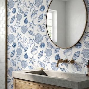 May include: A bathroom with a coastal theme. The walls feature white subway tiles and a wallpaper with blue seashell and starfish patterns. A wooden vanity with a concrete countertop and a round mirror complete the look. A shower with a brass showerhead is visible.