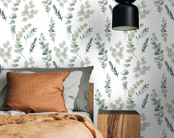 May include: A bedroom scene featuring a bed with an orange pillow and a tan blanket. The wall is decorated with a white wallpaper with a repeating pattern of green and blue eucalyptus leaves. A black pendant light hangs from the ceiling.