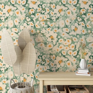 Op de afbeelding: Een behang met een witte achtergrond en een groen en geel bloemenpatroon. Het behang is getoond op een muur achter een houten tafel met een vaas met gedroogde bladeren en een stapel boeken.