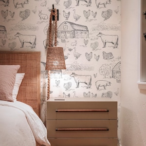 May include: A bedroom scene with a light-coloured bedside table with three drawers and brown handles. A woven lampshade hangs above the table. The wallpaper has a farm animal pattern. A bed with a woven headboard is visible.