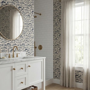 May include: A bathroom with a white vanity, a round gold mirror, and a shower. The walls are decorated with wallpaper featuring a pattern of blue and gray fish. The shower has white subway tiles and gold fixtures.