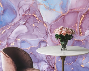 May include: A white marble table with a gold base sits in front of a purple and blue abstract wall with gold accents. A bouquet of pink roses sits in a clear glass vase on the table. A plush, dark pink velvet chair sits to the left of the table.