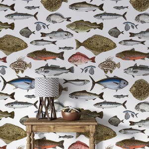 Könnte beinhalten: Ein weißer Hintergrund mit einem sich wiederholenden Muster aus verschiedenen Fischarten, darunter Thunfisch, Kabeljau und Flunder. Die Fische sind im Vintage-Stil mit detaillierten Linien und Schattierungen gezeichnet. Eine Lampe mit einem grauen und weißen Chevron-Muster-Schirm steht auf einem Holztisch vor der Tapete.