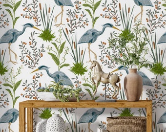 May include: A wooden console table with a white background featuring a repeating pattern of blue herons, green reeds, and orange berries. The table has two shelves and is decorated with a white ceramic vase, a white ceramic vase with cutouts, a wicker basket, a stack of books, and a decorative horse figurine.