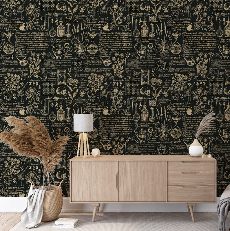 May include: A black and gold wallpaper with a repeating pattern of botanical illustrations, including flowers, plants, and herbs. The pattern also includes various symbols, such as stars, moons, and hands. The wallpaper is displayed on a wall in a room with a wooden dresser, a lamp, and a vase with dried flowers.
