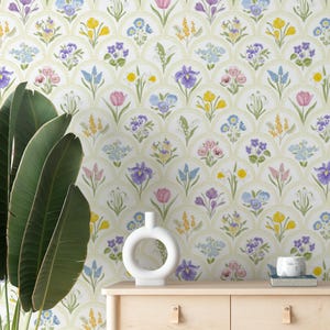 May include: A floral wallpaper design featuring a repeating pattern of colorful flowers in shades of purple, pink, yellow, and blue. A white vase with a circular design sits on a wooden cabinet. A large green plant is on the left.