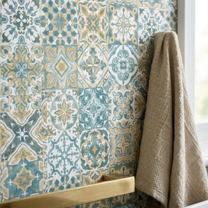 May include: A wall covered in square tiles featuring a repeating pattern of teal, white, and mustard yellow geometric and floral motifs. A horizontal brushed gold metal towel bar is mounted on the wall, with a beige textured towel hanging nearby.