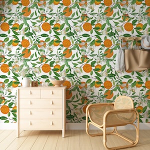 Op de afbeelding: Een kamer met een witte muur bedekt met een herhalend patroon van sinaasappels, groene bladeren en witte bloemen. Een lichte houten dressoir, een rieten stoel en andere decoraties bevinden zich in de kamer.