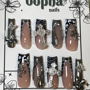 Puede incluir: Conjunto de diez uñas postizas con un diseño de estampado de cebra en blanco y negro. Cada uña presenta una cruz plateada con pedrería. Las uñas se muestran en una tarjeta blanca con el texto "bopha nails" y "HANDPAINTED WITH LOVE BY:".