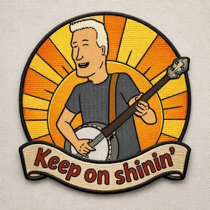 May include: Embroidered patch featuring a cartoon man playing a banjo against a yellow and orange sunburst background. The patch includes the text "Keep on shinin'" in a banner below the figure. The design has a vintage, country music aesthetic.