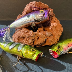 May include: Three fishing lures with different color patterns and feather tails. The lures are silver with purple accents, green with gold accents, and green with pink accents.