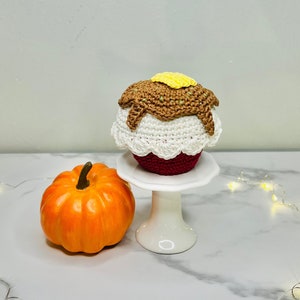 以下が含まれることがあります： 白いケーキスタンドに、茶色のフロスティングと黄色のバターが乗った、かぎ針編みのカップケーキが飾られています。白い大理石の表面に、小さなオレンジ色のカボチャと白いフェアリーライトの紐が置かれています。