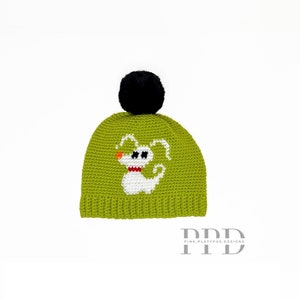 Op de afbeelding: Groene gebreide muts met een zwarte pompon en een wit geborduurd spookontwerp. De muts heeft de tekst "PPD PINK.PLATYPUS.DESIGNS" aan de onderkant.